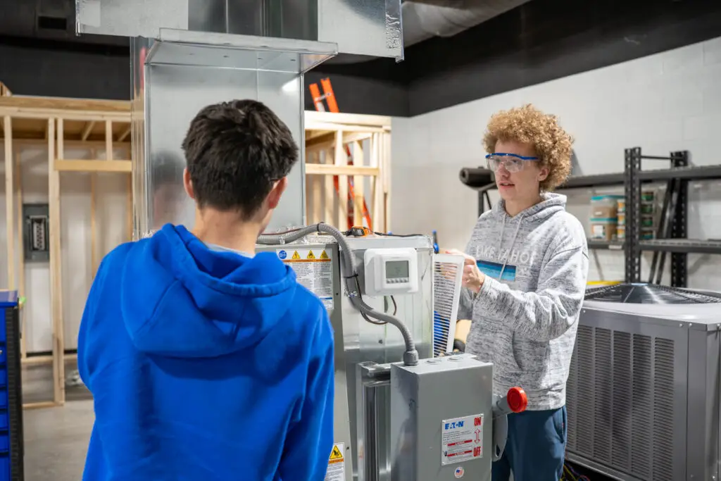 Two young men perform maintenance on an air conditioning unit in the high school trades lab.